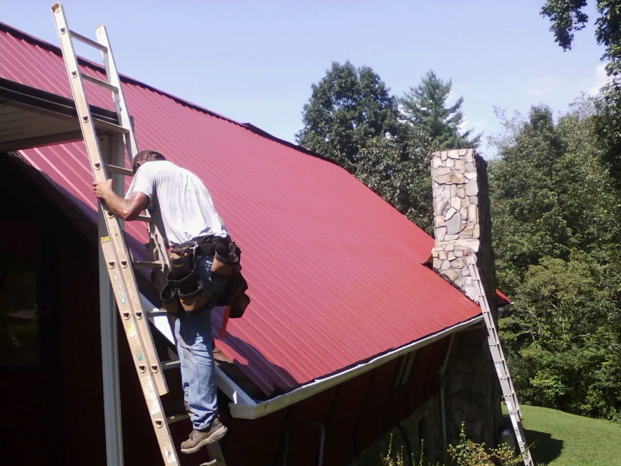 Roofer on a ladder beside a red metal roof and stone chimney — Soffit & Fascia Repair by Roofing Brothers in Fred